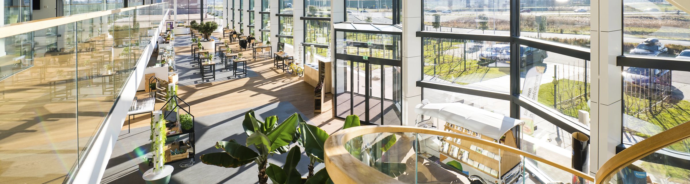 Interieur van het Van Gelder Proefcentrum: atrium met houten tafels, planten en spiraaltrap achter de glazen facade